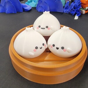 Peut inclure: Trois figurines de raviolis blancs souriants, avec des joues roses et des yeux noirs, sont pos&eacute;es dans un panier vapeur rond marron. Les raviolis ont un design mignon et cartoon. Le panier a une texture c&ocirc;tel&eacute;e.