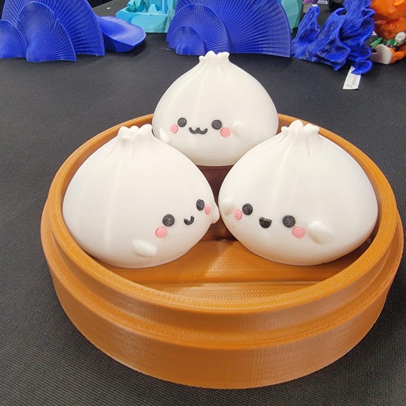 Peut inclure: Trois figurines de raviolis blancs souriants, avec des joues roses et des yeux noirs, sont pos&eacute;es dans un panier vapeur rond marron. Les raviolis ont un design mignon et cartoon. Le panier a une texture c&ocirc;tel&eacute;e.