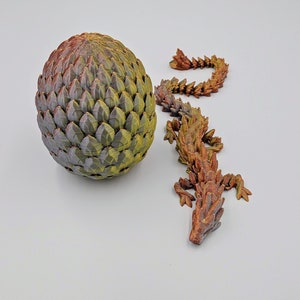 May include: A green and brown dragon egg with a small, brown, scaly dragon figurine next to it.