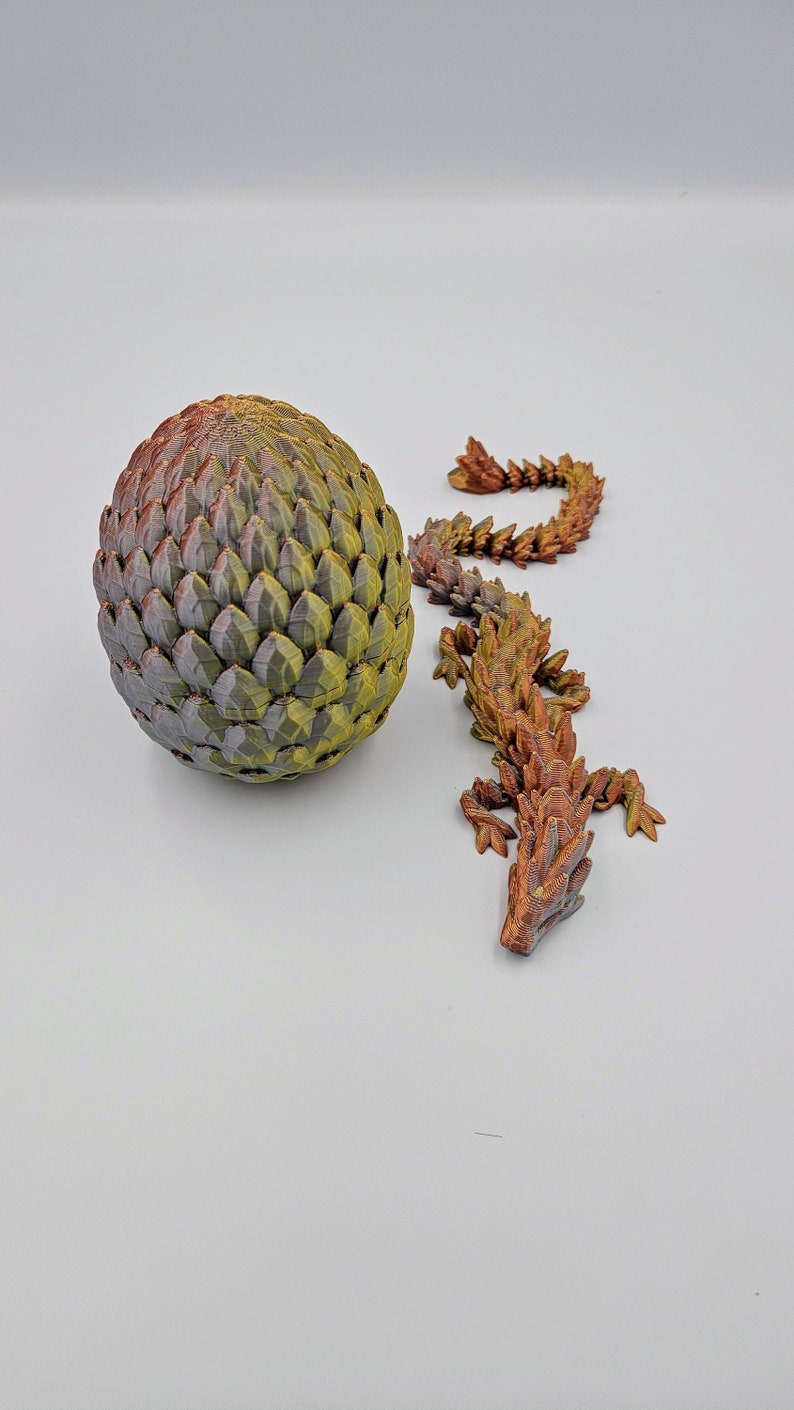 May include: A green and brown dragon egg with a small, brown, scaly dragon figurine next to it.