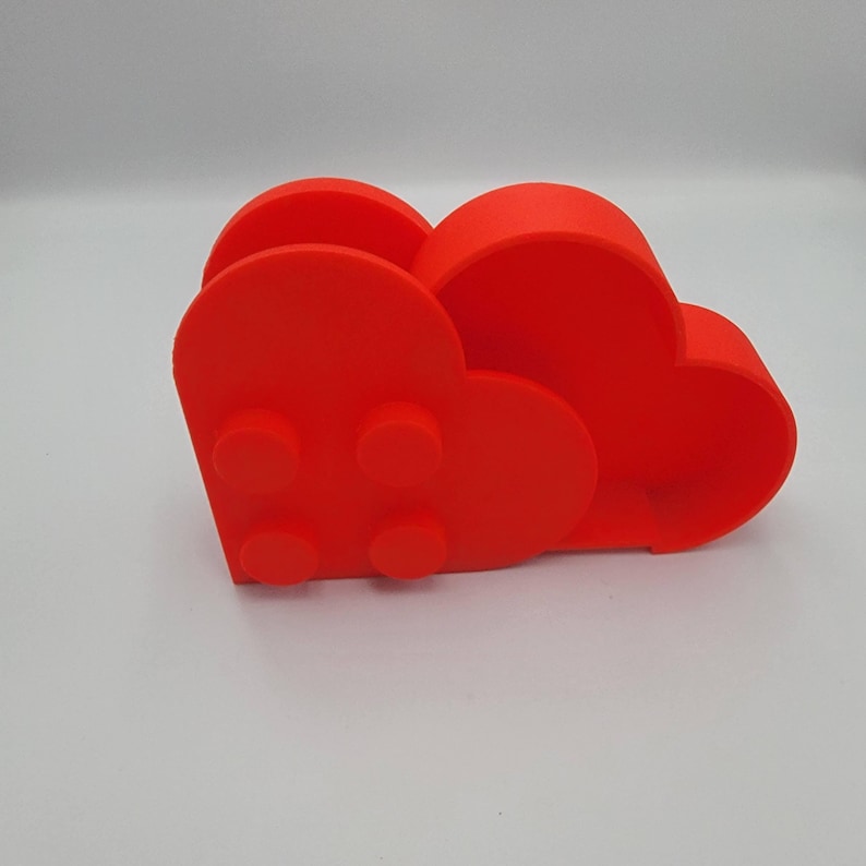 May include: A bright red, 3D-printed desk organizer shaped like a cloud. The organizer has a heart-shaped design with four circular studs on the front, resembling a building block toy. The organizer is made of a smooth, solid material.