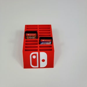 Może przedstawiać: Czerwony plastikowy stojak na karty do gier Nintendo Switch z białym logo Nintendo Switch. Stojak ma 24 gniazda na karty do gier, a w dw&oacute;ch gniazdach znajdują się karty. Karty są do gier "The Legend of Zelda: Breath of the Wild" i "Super Mario Odyssey".