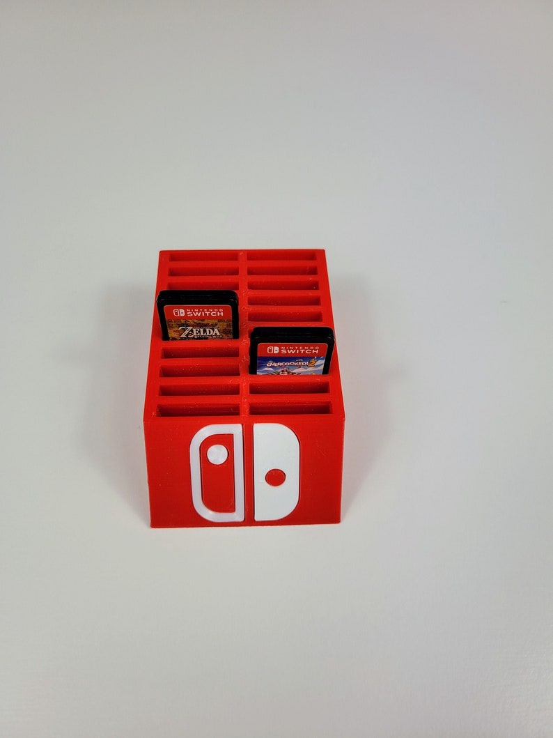Może przedstawiać: Czerwony plastikowy stojak na karty do gier Nintendo Switch z białym logo Nintendo Switch. Stojak ma 24 gniazda na karty do gier, a w dw&oacute;ch gniazdach znajdują się karty. Karty są do gier "The Legend of Zelda: Breath of the Wild" i "Super Mario Odyssey".