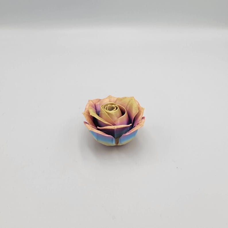 May include: A multi-colored rose with a metallic finish. The petals transition from yellow to pink and blue, creating a vibrant, iridescent effect. The rose is a decorative item, possibly a sculpture or a unique craft piece.
