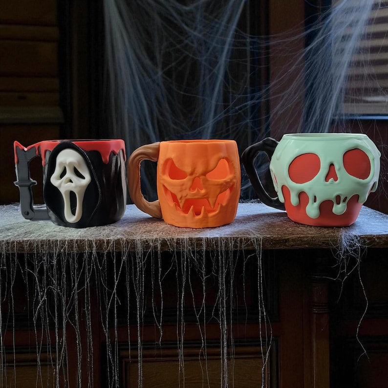 Pode incluir: Tr&ecirc;s canecas com tema de Halloween. Uma apresenta um design de Ceifador negro com uma m&aacute;scara branca, outra &eacute; uma ab&oacute;bora laranja e a terceira &eacute; uma caneca vermelha com um design de caveira verde. Todas as canecas t&ecirc;m al&ccedil;as.