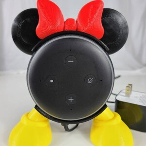 Puede incluir: Un soporte impreso en 3D para un Amazon Echo Dot, dise&ntilde;ado para parecerse a Mickey Mouse. El soporte tiene orejas rojas con un lazo rojo, pies amarillos y un altavoz negro con botones.