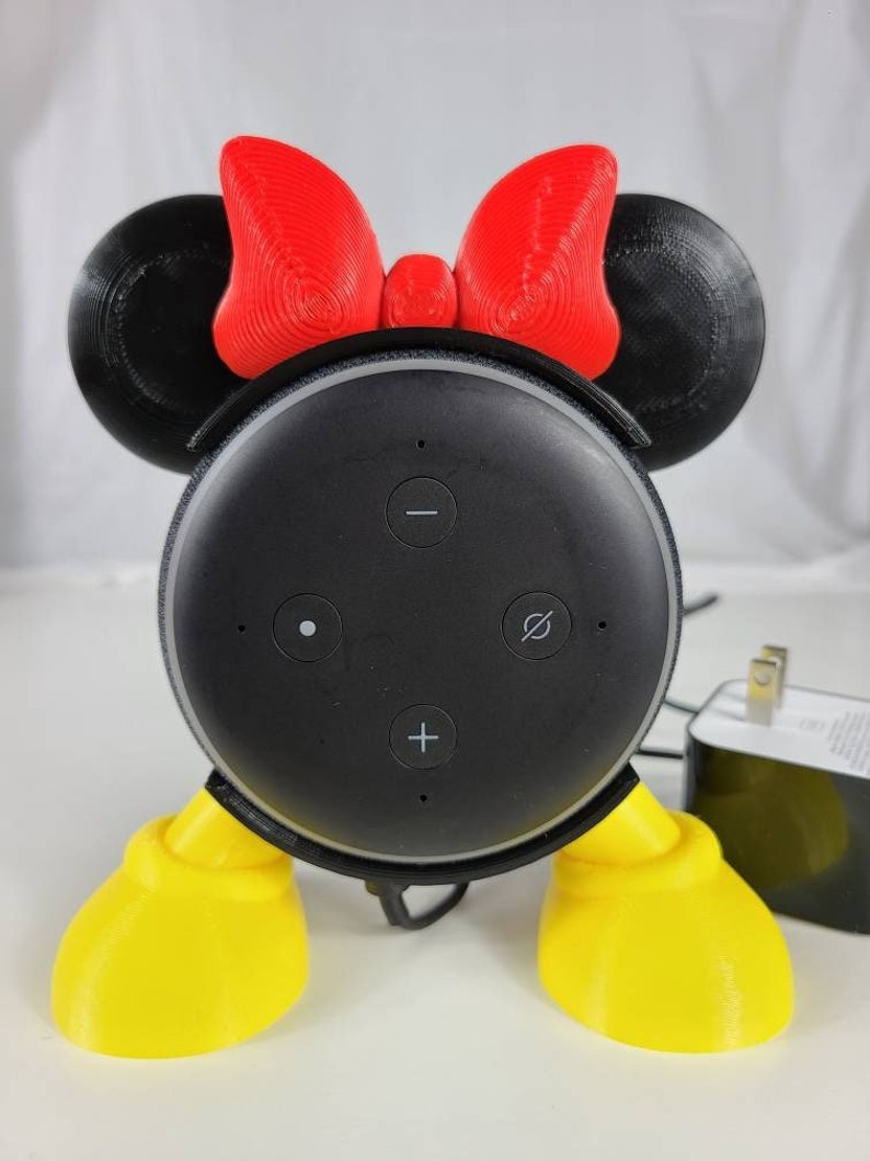Puede incluir: Un soporte impreso en 3D para un Amazon Echo Dot, dise&ntilde;ado para parecerse a Mickey Mouse. El soporte tiene orejas rojas con un lazo rojo, pies amarillos y un altavoz negro con botones.