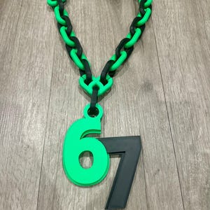 May include: A chunky necklace with a green and black chain design. A green number 6 is attached to the chain, along with a black number 7. The necklace is displayed on a wooden surface.