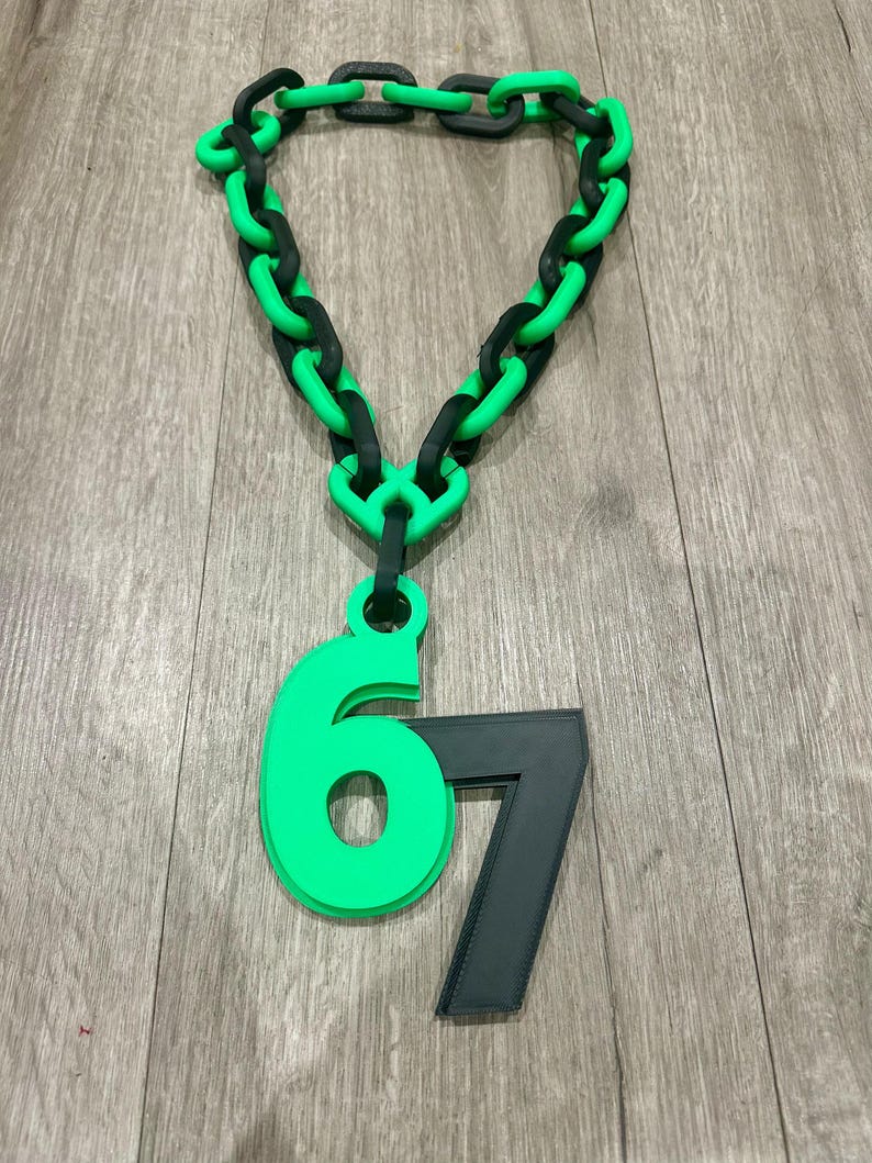 May include: A chunky necklace with a green and black chain design. A green number 6 is attached to the chain, along with a black number 7. The necklace is displayed on a wooden surface.