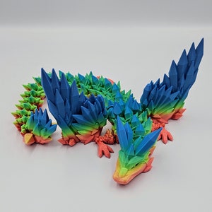 May include: A rainbow-coloured 3D printed dragon figurine with a spiky, crystal-like design. The dragon has wings, a long tail, and a pointed snout.