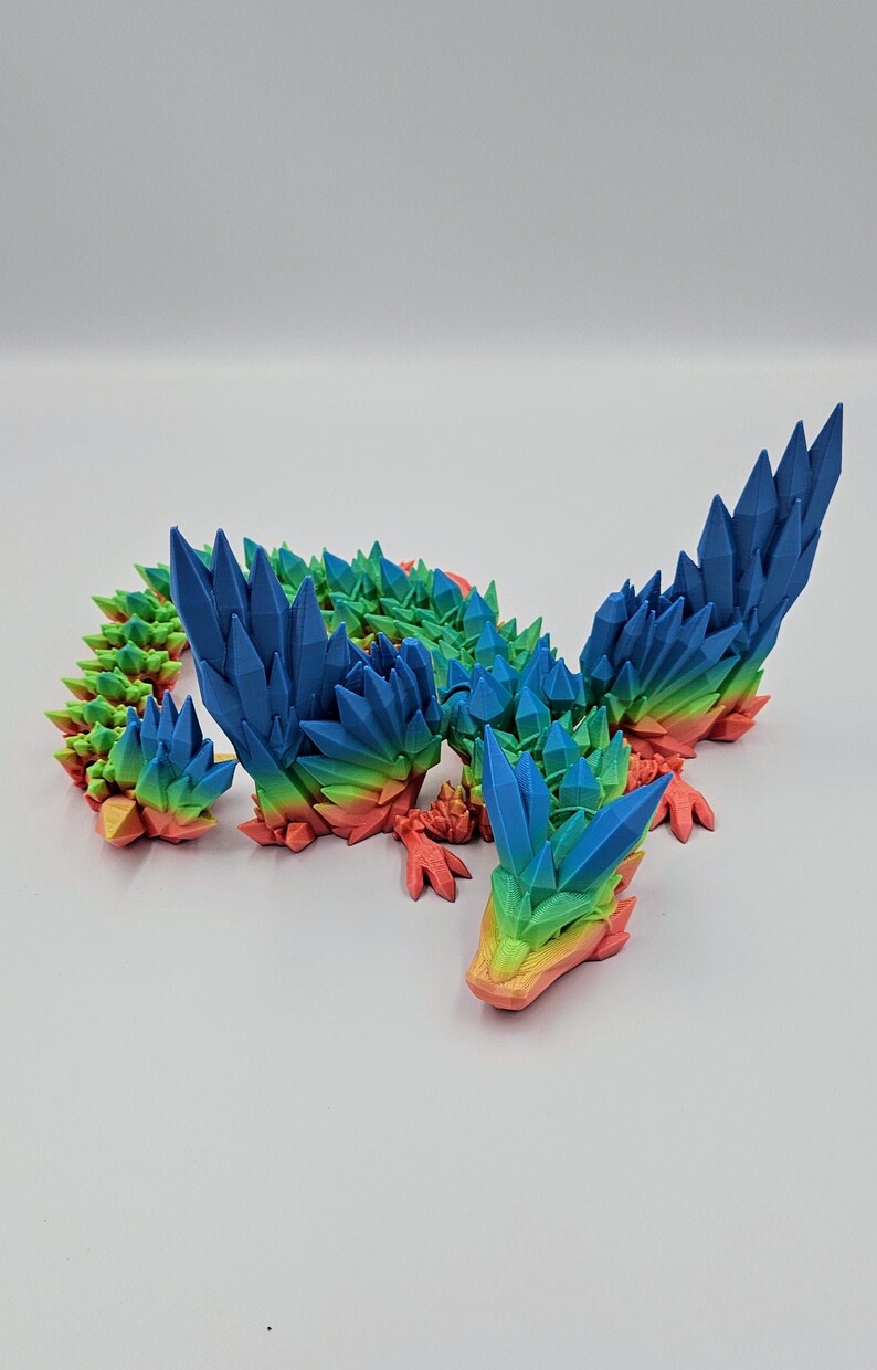 May include: A rainbow-coloured 3D printed dragon figurine with a spiky, crystal-like design. The dragon has wings, a long tail, and a pointed snout.