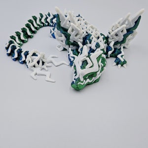 May include: A 3D printed dragon figurine with a white and green colour scheme. The dragon has a long, segmented tail and a detailed head with sharp teeth.