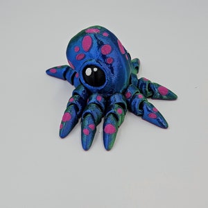 Puede incluir: Una peque&ntilde;a figura de pulpo azul y verde con manchas rosas. El pulpo tiene un ojo grande y negro y ocho tent&aacute;culos.