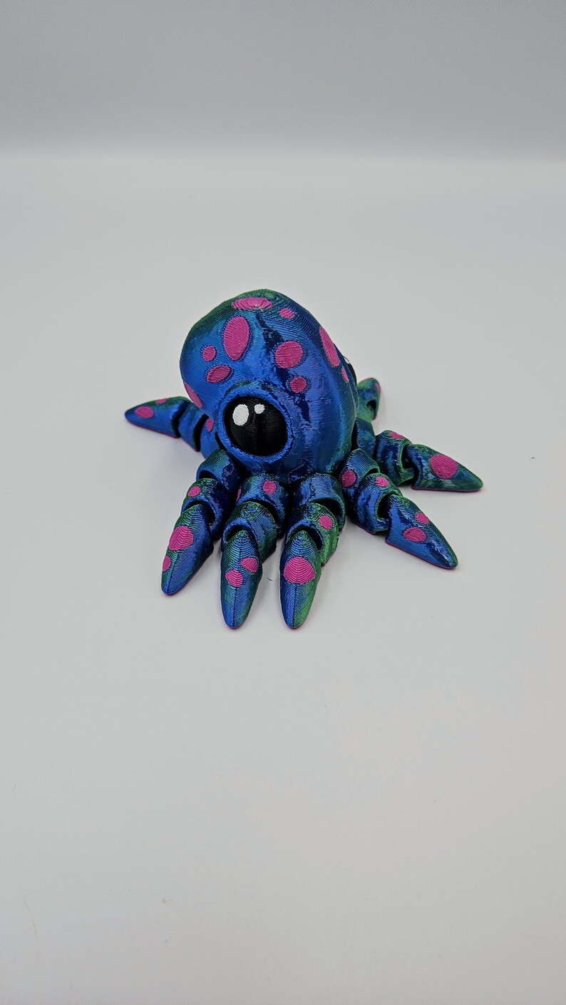 Puede incluir: Una peque&ntilde;a figura de pulpo azul y verde con manchas rosas. El pulpo tiene un ojo grande y negro y ocho tent&aacute;culos.