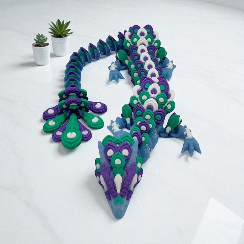 May include: A detailed, articulated dragon sculpture in shades of blue, green, purple, and white. The dragon is posed on a white surface, with a decorative tail and head. Two small potted succulents are in the background.