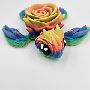 May include: A rainbow-colored 3D printed turtle with a rose on its back. The turtle has large black eyes and a detailed shell.