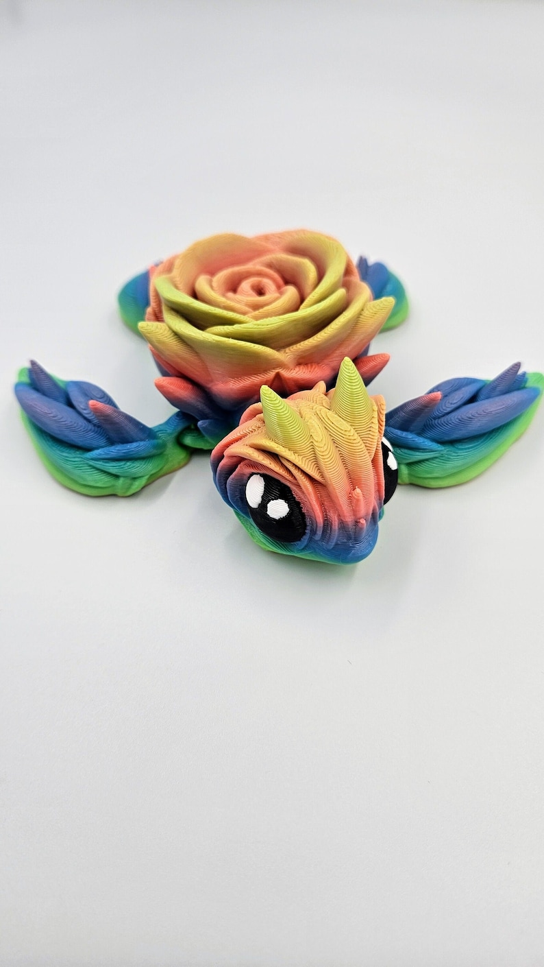 May include: A rainbow-colored 3D printed turtle with a rose on its back. The turtle has large black eyes and a detailed shell.