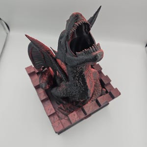 May include: A black and red 3D-printed dinosaur sculpture with an open mouth, sharp teeth, and large wings. The sculpture is mounted on a red brick-like base. The dinosaur's head is tilted back, and its mouth is wide open, showcasing its teeth.