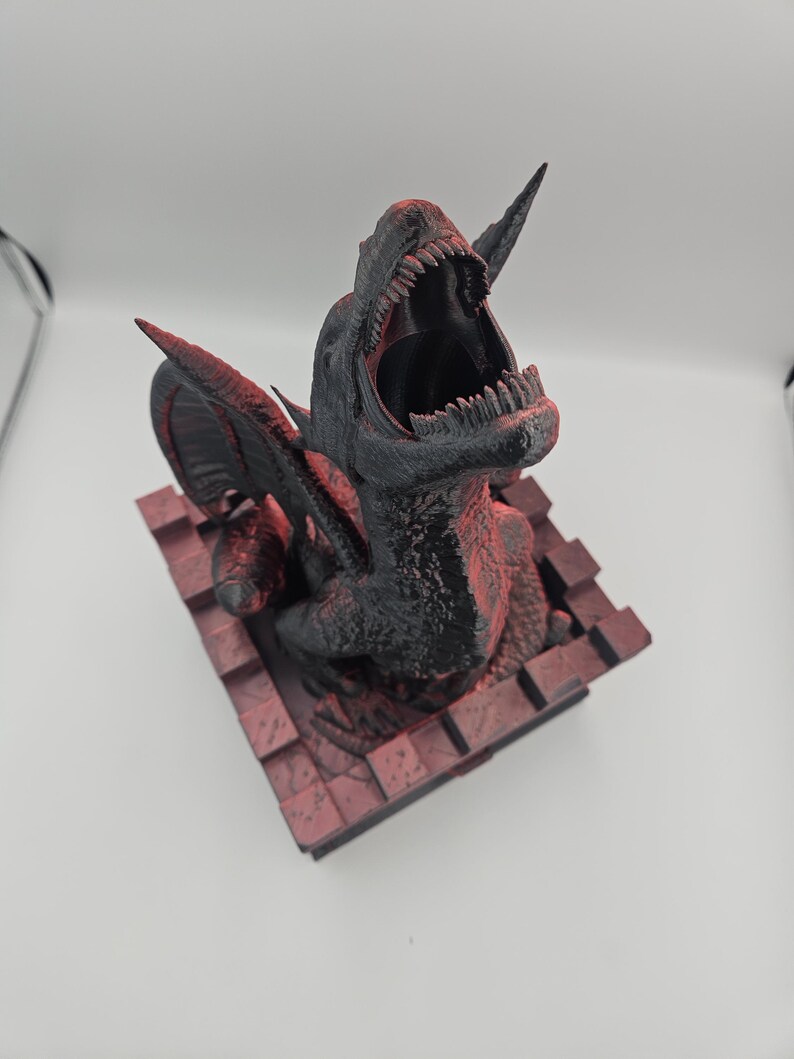 May include: A black and red 3D-printed dinosaur sculpture with an open mouth, sharp teeth, and large wings. The sculpture is mounted on a red brick-like base. The dinosaur's head is tilted back, and its mouth is wide open, showcasing its teeth.