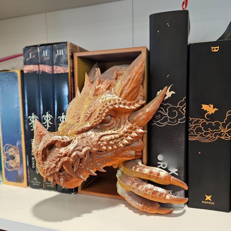 May include: A detailed, bronze-colored dragon head bookend. The dragon's head is emerging from a wooden box, with sharp teeth and textured scales. The bookend is designed to hold books on a shelf, adding a fantasy theme to a bookshelf.