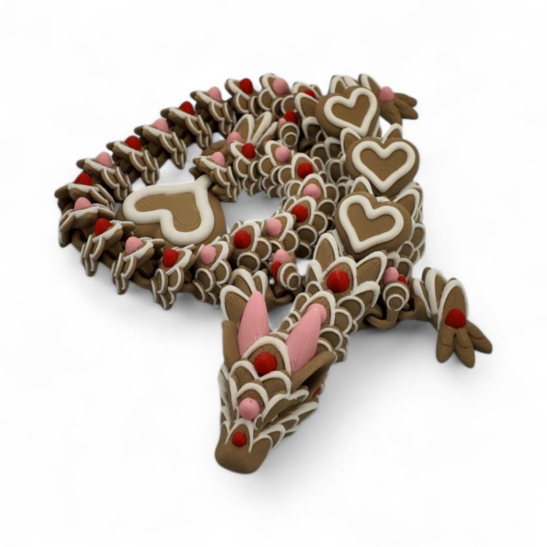 May include: A brown gingerbread dragon with white icing details, pink accents, and red candies. The dragon's body is made of interconnected gingerbread pieces, with heart-shaped cookies and a detailed head.