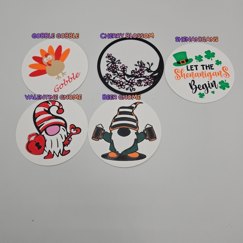 May include: A collection of five circular decorative coasters. Designs include a turkey with "Gobble Gobble", a cherry blossom, a gnome with beer mugs, a gnome with a heart and key, and a green hat with "Let the Shenanigans Begin".