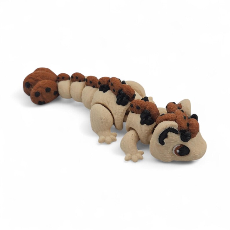 May include: A tan and brown articulated toy lizard with a segmented body and a smiling face. The toy has brown spots and accents, and the tail is made of several brown, rounded segments. The lizard is posed on a white background.