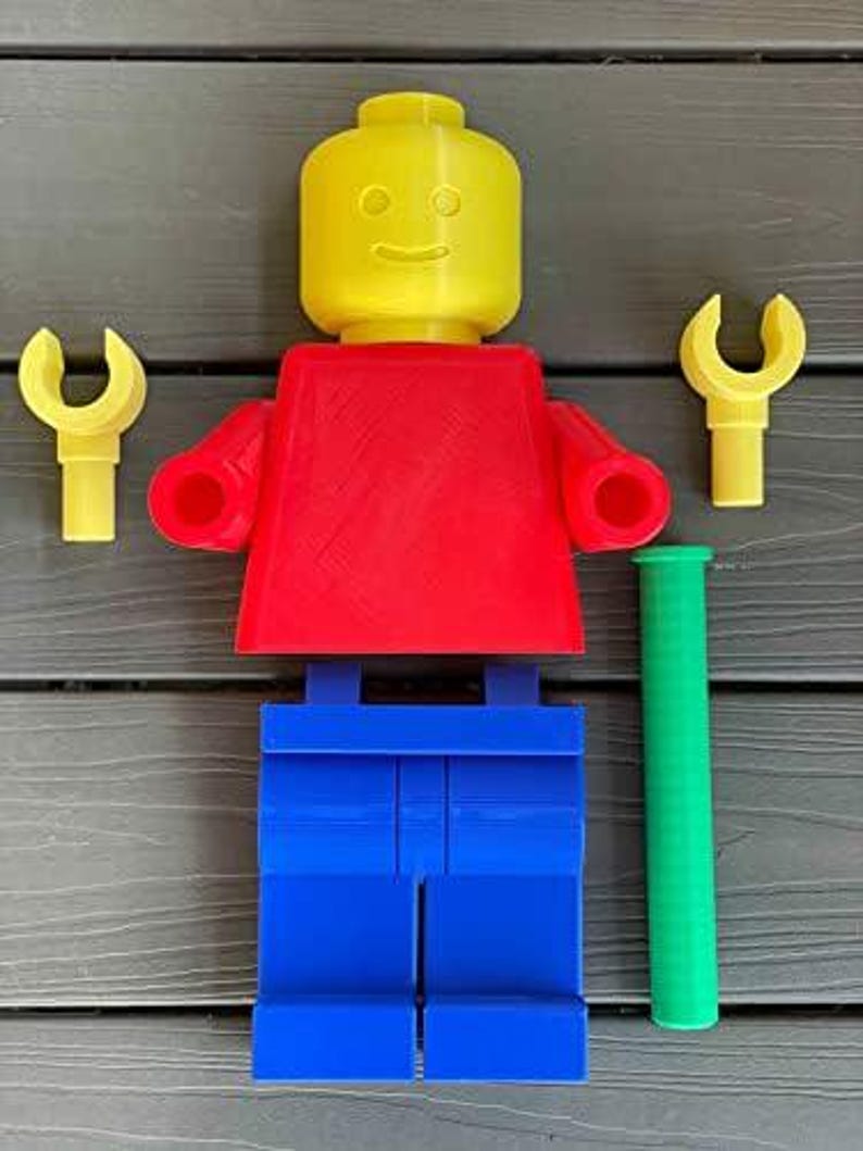 K&ouml;nnte beinhalten: Eine gro&szlig;e, farbenfrohe Lego-Figur mit gelbem Kopf, rotem Torso und blauen Beinen. Zwei gelbe schraubenschl&uuml;sself&ouml;rmige Zubeh&ouml;rteile und ein gr&uuml;ner Zylinder sind in der N&auml;he der Figur platziert. Die Figur ist ein lustiges, &uuml;bergro&szlig;es Spielzeug.