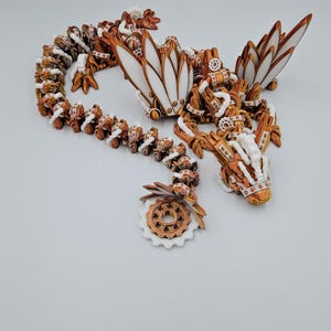 May include: A white and copper-coloured 3D printed dragon with intricate details and a gear-shaped tail. The dragon has wings and a long, winding body.