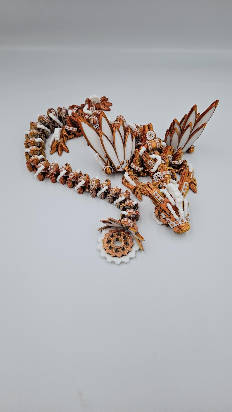 May include: A white and copper-coloured 3D printed dragon with intricate details and a gear-shaped tail. The dragon has wings and a long, winding body.