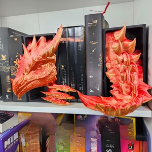 May include: A pair of dragon-themed bookends in shades of red and gold. The bookends are designed to resemble a dragon's head and tail, adding a fantasy element to a bookshelf. The dragon's head is on the left, and the tail is on the right.