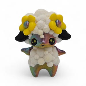 May include: A whimsical 3D-printed figurine of a sheep-like creature. It features a fluffy white body, a rainbow-colored face and legs, and large, expressive eyes. The sheep wears a crown of yellow flowers and has black ears.