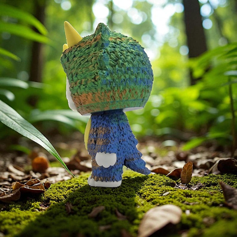 K&ouml;nnte beinhalten: Eine farbenfrohe Figur einer Kreatur mit einem strukturierten, gr&uuml;n-blauen Kopf, einem gelben Horn und einem blauen K&ouml;rper. Die Figur steht auf einem Bett aus gr&uuml;nem Moos in einer Waldlandschaft.