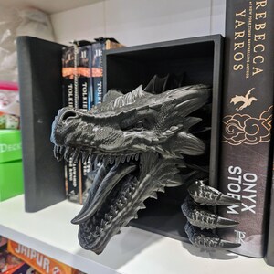 Pode incluir: Um suporte de livros de drag&atilde;o preto com escamas detalhadas e boca aberta, posicionado entre livros numa prateleira. A cabe&ccedil;a e as garras do drag&atilde;o s&atilde;o vis&iacute;veis, adicionando um elemento de fantasia. Livros de Tolkien e Rebecca Yarros s&atilde;o vis&iacute;veis.