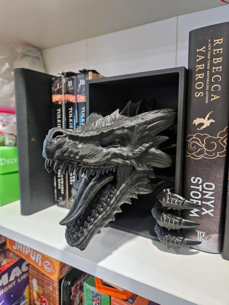 Pode incluir: Um suporte de livros de drag&atilde;o preto com escamas detalhadas e boca aberta, posicionado entre livros numa prateleira. A cabe&ccedil;a e as garras do drag&atilde;o s&atilde;o vis&iacute;veis, adicionando um elemento de fantasia. Livros de Tolkien e Rebecca Yarros s&atilde;o vis&iacute;veis.