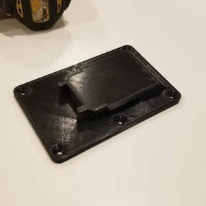 May include: A black 3D printed wall mount for a power tool battery. The mount has a slot for the battery and four screw holes.