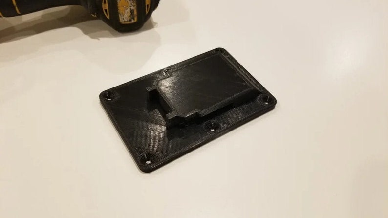 May include: A black 3D printed wall mount for a power tool battery. The mount has a slot for the battery and four screw holes.