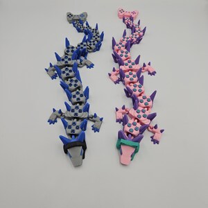 May include: Two articulated dragon toys, one gray and blue, the other pink and purple. Each dragon is constructed from interlocking game controller-shaped segments, with spiky protrusions along the spine and head. The toys are displayed on a white surface.