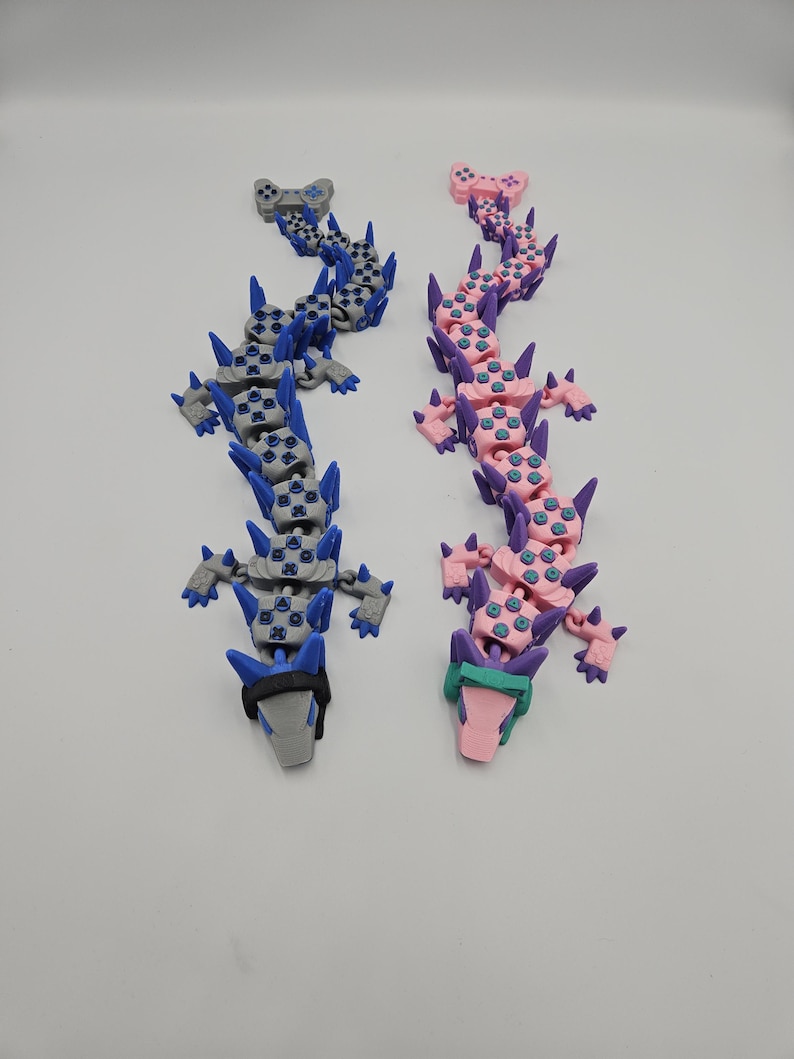 May include: Two articulated dragon toys, one gray and blue, the other pink and purple. Each dragon is constructed from interlocking game controller-shaped segments, with spiky protrusions along the spine and head. The toys are displayed on a white surface.