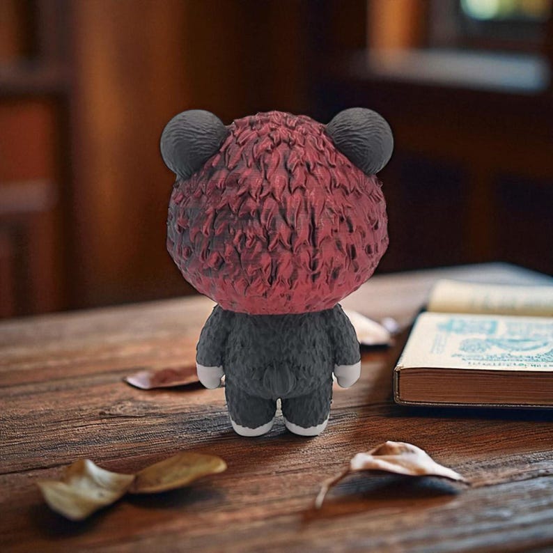 May include: A small, stylized bear figurine with a textured, burgundy head and dark gray body. The bear has small, round black ears and white feet. It is positioned on a wooden surface with scattered leaves and a book in the background.