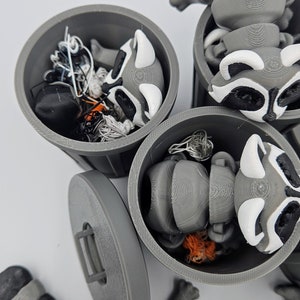 May include: 3D-printed raccoon figurines in gray, white, and black, with articulated bodies and trash can containers. The raccoons have detailed features, including black masks and white markings. The trash cans are gray with lids, and some contain small items.