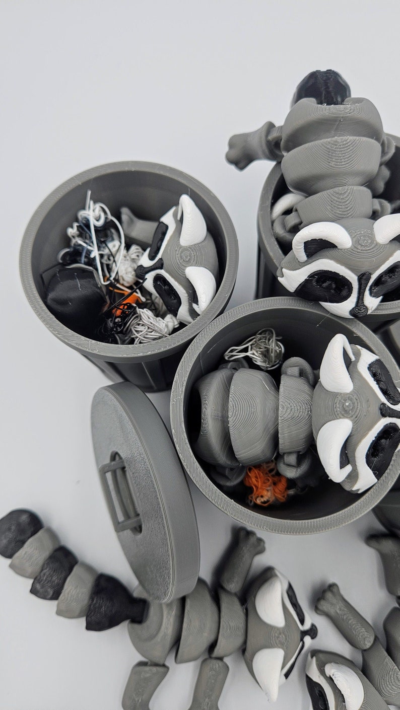 May include: 3D-printed raccoon figurines in gray, white, and black, with articulated bodies and trash can containers. The raccoons have detailed features, including black masks and white markings. The trash cans are gray with lids, and some contain small items.