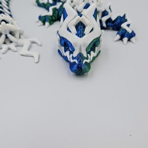 May include: A 3D printed dragon figurine with a white and blue body and green accents. The dragon is curled up in a coil shape.