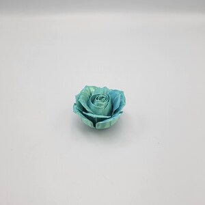 May include: A single, teal-colored rose with a gradient effect, transitioning from a lighter green to a deeper blue. The rose is in full bloom, with detailed petals and a realistic appearance. The flower is set against a plain white background.