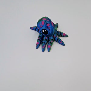 Puede incluir: Una peque&ntilde;a figura de pulpo azul con manchas rosas y detalles verdes. El pulpo tiene un solo ojo negro y ocho tent&aacute;culos.