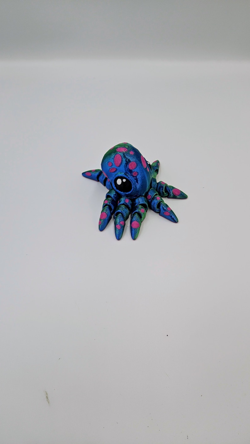 Puede incluir: Una peque&ntilde;a figura de pulpo azul con manchas rosas y detalles verdes. El pulpo tiene un solo ojo negro y ocho tent&aacute;culos.