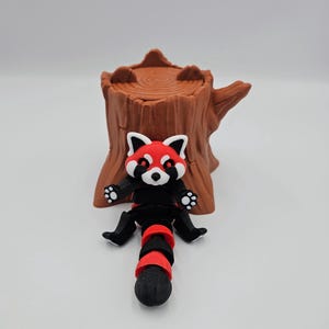 May include: A red panda figurine with black and red striped tail sitting on a brown tree stump. The figurine has a white face with black eyes and a red nose.