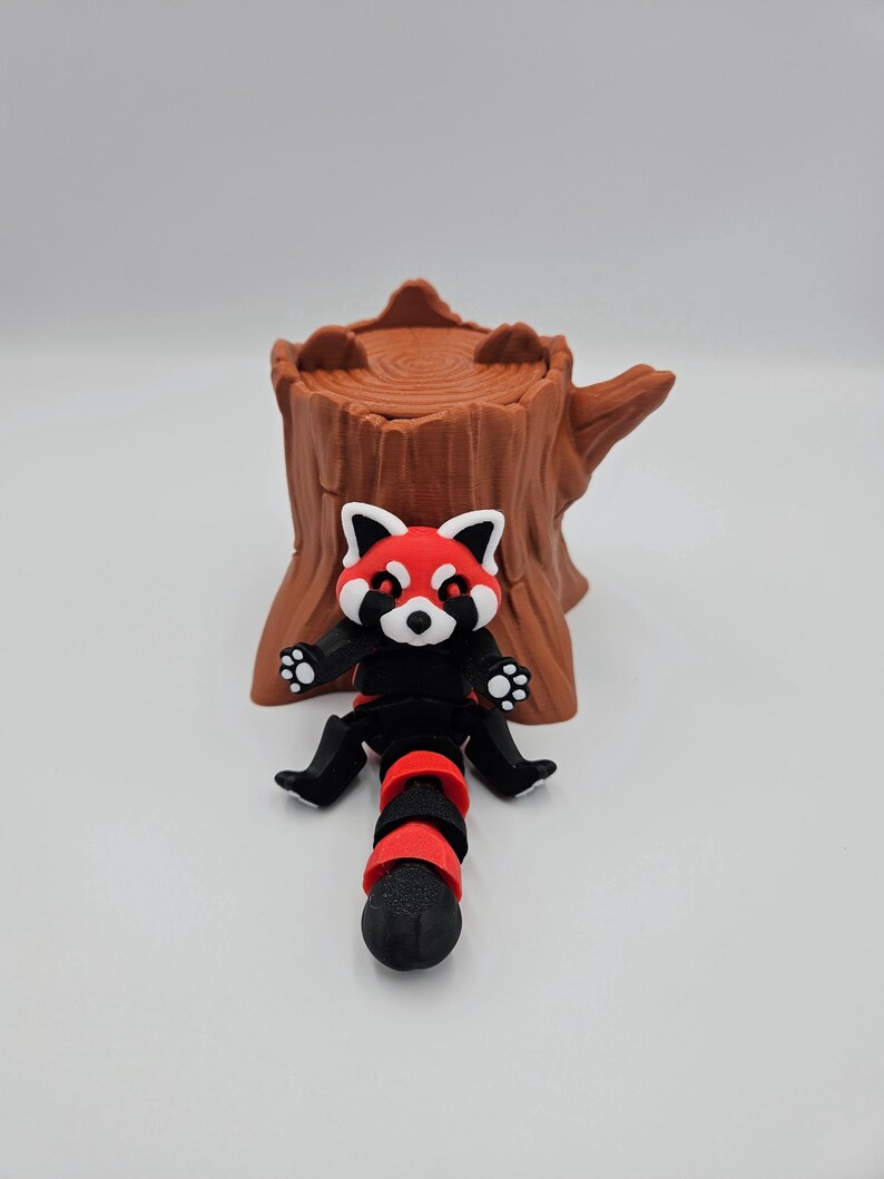 May include: A red panda figurine with black and red striped tail sitting on a brown tree stump. The figurine has a white face with black eyes and a red nose.