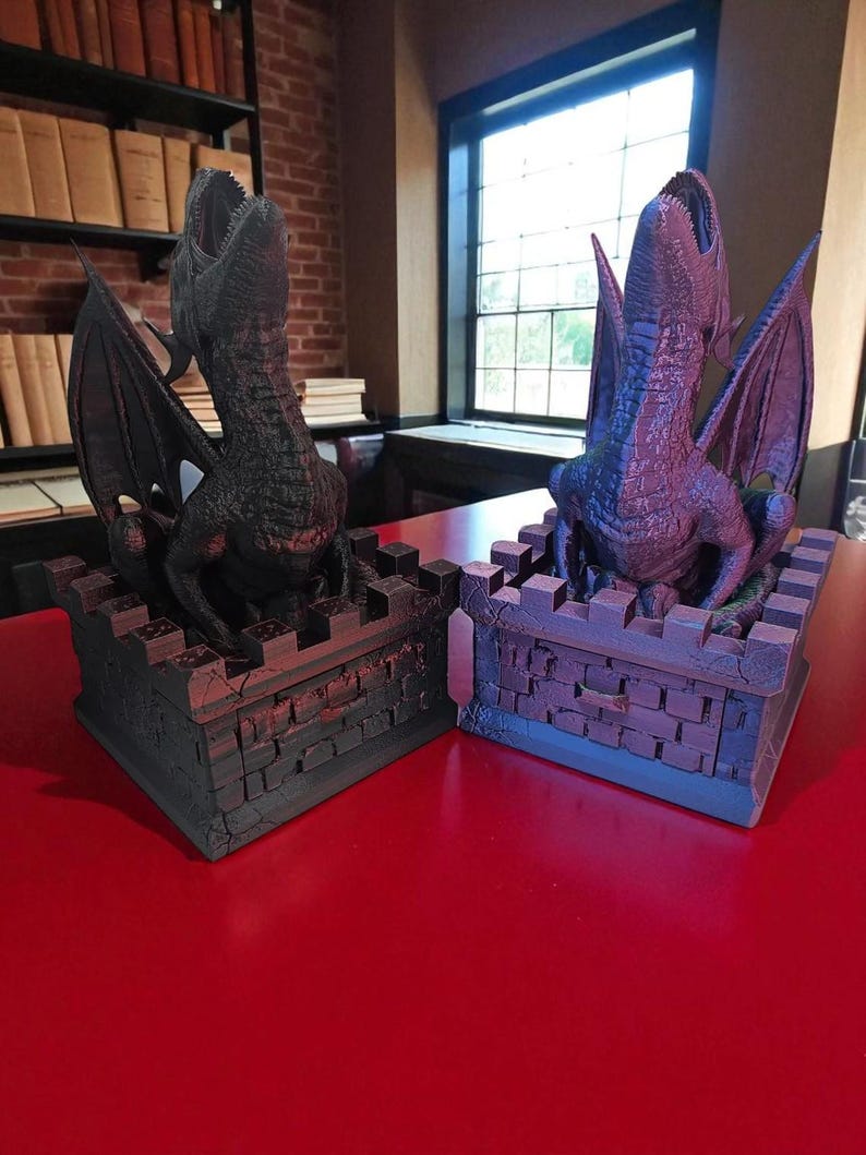May include: Two dragon bookends. Each dragon is perched atop a castle-like base with open mouths and outstretched wings. One is dark gray, the other is purple. The bookends are on a red surface, with a library background.