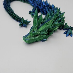 May include: A 3D printed dragon figurine with a green and blue iridescent finish. The dragon is curled up in a defensive posture with its head resting on its tail.